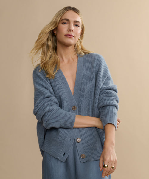 A woman with long blonde hair wears the Cropped Cashmere Cocoon Cardigan and matching pants, standing against a plain beige background and looking confidently at the camera.