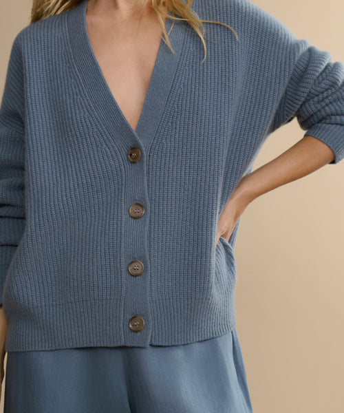 A person wears the Cropped Cashmere Cocoon Cardigan with large buttons and matching blue pants, standing with one hand in their pocket against a neutral background.