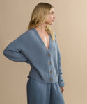 A woman with long blonde hair stands in profile against a beige background, wearing a loose, light blue Cropped Cashmere Cocoon Cardigan with buttons and matching wide-leg pants.
