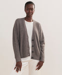 A person models the Cropped Cashmere Cocoon Cardigan in gray over a white shirt and pants, standing confidently against a plain light background.