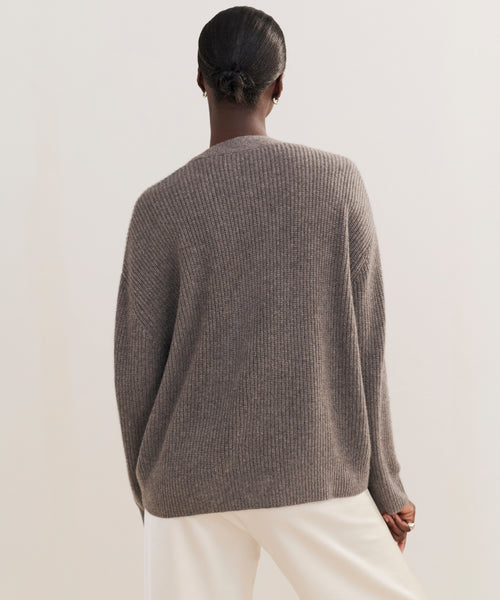 Seen from behind against a plain, light background, a person with dark hair in a low bun wears the Cropped Cashmere Cocoon Cardigan in gray with white pants.