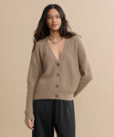 A woman with long dark hair wears the Cropped Cashmere Cocoon Cardigan in undyed stone with a deep V-neck and black pants, standing against a beige background.