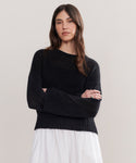 A woman with long brown hair stands with arms crossed, wearing the Cropped Linen Crewneck in black and a flowing white skirt, against a plain light-colored background.