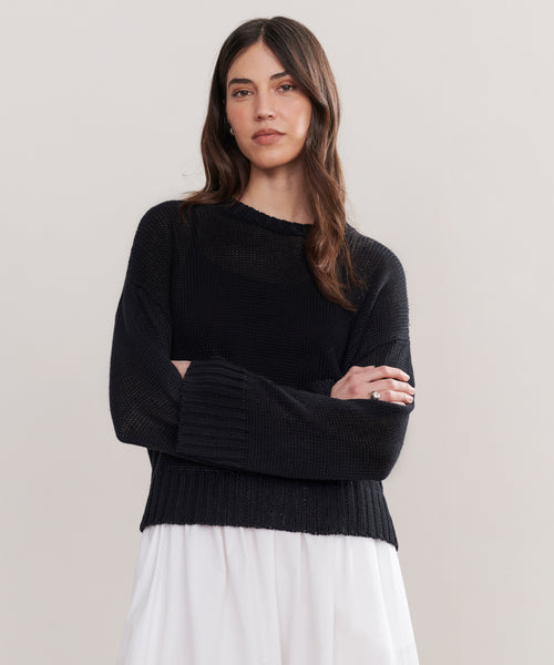 A woman with long brown hair stands with arms crossed, wearing the Cropped Linen Crewneck in black and a flowing white skirt, against a plain light-colored background.