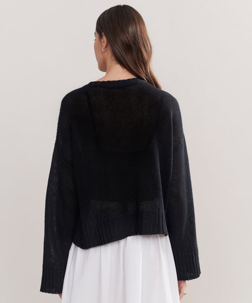 A woman with long brown hair, seen from behind, wears the Cropped Linen Crewneck—a relaxed-fit, slightly sheer black sweater—over a white dress. The long sleeves and loose drape add effortless style against a light background.