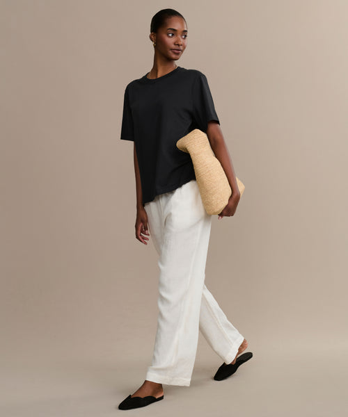 Wearing a relaxed fit Crosby Tee, white pants, and black slippers, a woman walks against a neutral background holding a textured beige pillow under her arm.