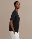 A woman stands against a beige background, wearing the black Crosby Tee in a relaxed fit and white pants. She faces sideways, hands in her pockets, looking at the camera with a gentle smile.