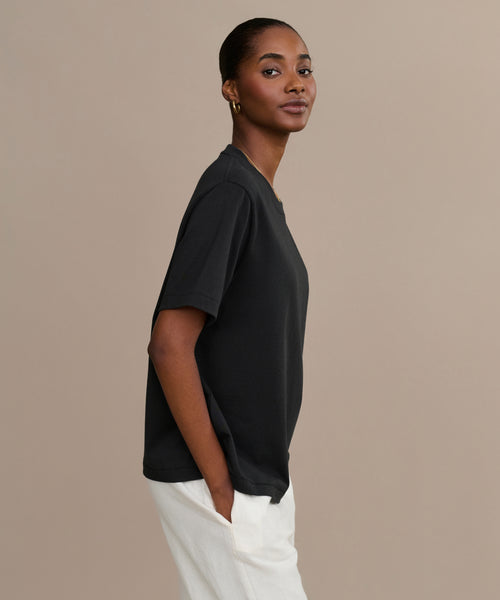 A woman stands against a beige background, wearing the black Crosby Tee in a relaxed fit and white pants. She faces sideways, hands in her pockets, looking at the camera with a gentle smile.
