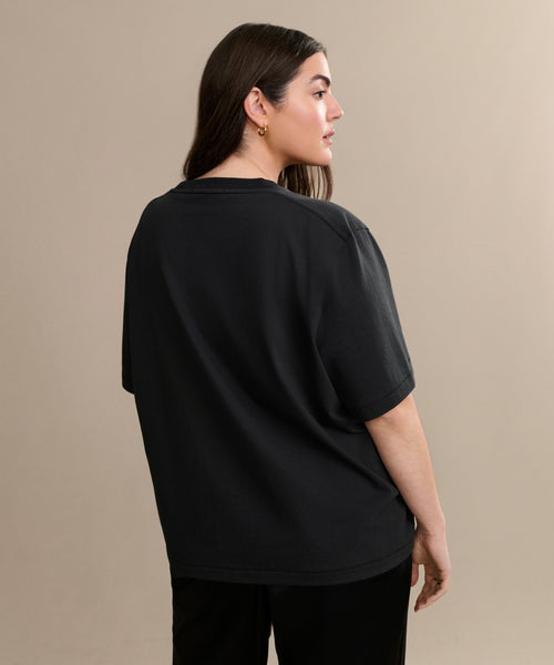 A woman with long brown hair wears the Crosby Tee, a relaxed fit top in premium jersey cotton, paired with black pants. She stands with her back turned, looking to the side against a plain beige background.