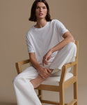 Woman sitting on a wooden chair wearing a white outfit against a beige background