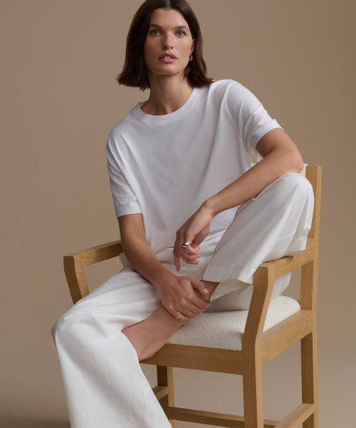 Woman sitting on a wooden chair wearing a white outfit against a beige background