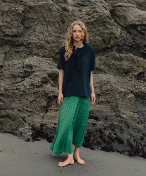 Woman in a dark top and green skirt standing against a rocky beach background