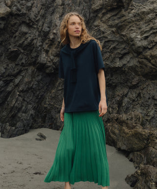 Woman wearing a dark blue top and green pleated skirt standing against a rocky background.