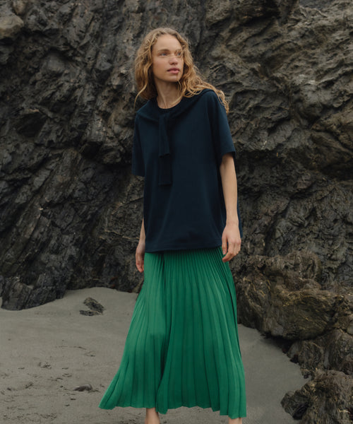 Woman wearing a dark blue top and green pleated skirt standing against a rocky background.