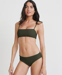 A woman with long dark hair poses against a plain white background, wearing the Cruz Bay Bikini Bottom in olive green, styled as a full-coverage bikini with a straight neckline and thin shoulder straps.