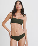 A woman with long dark hair stands against a white background, confidently modeling the Cruz Bay Bikini Bottom in olive green with thin straps and a high-cut design, one hand on her hip.