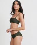 A woman with long dark hair models the Cruz Bay Bikini Bottom in olive green, standing sideways and looking at the camera against a plain white background.