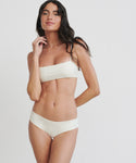 A woman with long dark hair poses against a plain background, wearing a minimalist, light-colored bikini with a straight bandeau top and the Cruz Bay Bikini Bottom, crafted from recycled nylon swimwear.