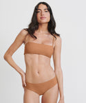 A woman with long dark hair models the Cruz Bay Bikini Bottom in light brown recycled nylon. She stands against a plain white background, looking at the camera with a relaxed expression, one hand on her hip.