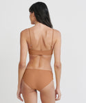 A woman with long dark hair stands with her back to the camera, wearing the Cruz Bay Bikini Bottom in light brown recycled nylon against a plain white background.