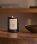 A Currant Glass Candle sits on a wooden table beside a shallow dish. Hand-poured in Los Angeles with natural coconut soy wax, it fills the room with a softly blurred, warm, and cozy atmosphere.