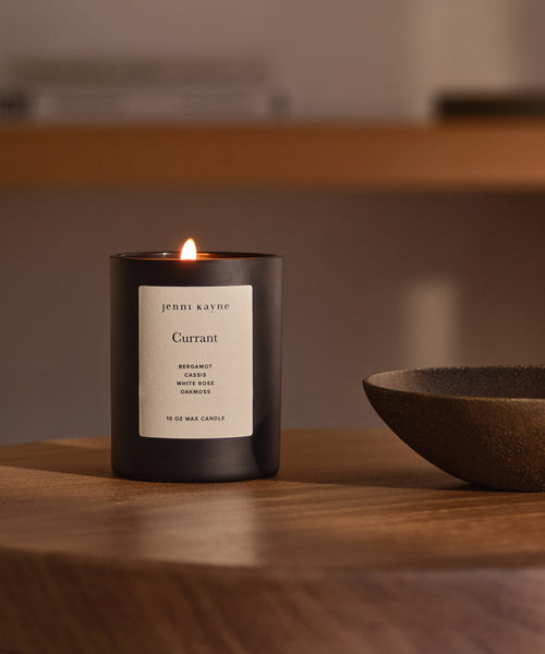 A Currant Glass Candle sits on a wooden table beside a shallow dish. Hand-poured in Los Angeles with natural coconut soy wax, it fills the room with a softly blurred, warm, and cozy atmosphere.