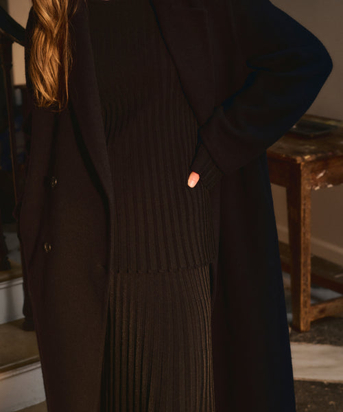 A person with long light brown hair stands indoors, one hand on their hip, wearing the Dara Top and a black coat. Their face is not visible.