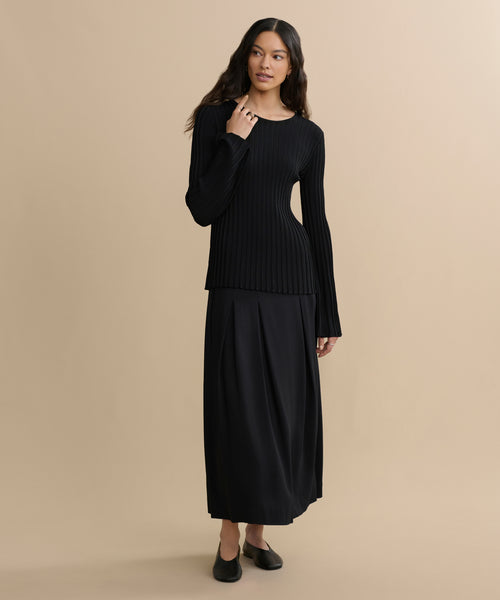 A woman wearing the Dara Top and a long black skirt stands against a beige background. She has long wavy hair, is wearing black flats, and has one hand touching her neck.