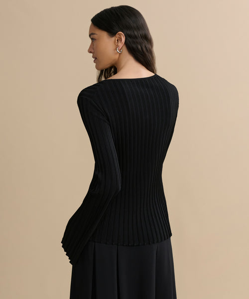 A woman with long, wavy dark hair stands facing away, wearing the fitted black Dara Top and a matching skirt against a plain beige background.