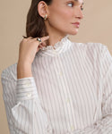 A woman models the Darcy Shirt—an ivory pinstripe top with a ruffled collar—styled with hoop earrings and rings, set against a beige backdrop.