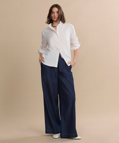 A woman stands against a beige background, wearing the ivory pinstripe Darcy Shirt, wide-leg dark blue jeans, and white shoes. With her hands in her pockets, she exudes a relaxed and confident vibe.