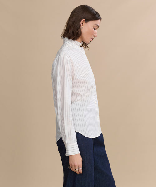 A woman with shoulder-length brown hair stands in profile against a beige background, wearing the Darcy Shirt—an ivory pinstripe shirt with a collared neckline—paired with dark blue pants.