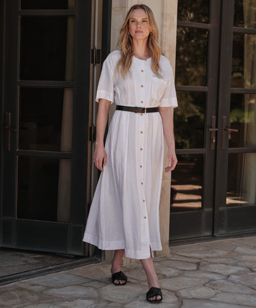 A person stands on a sunlit stone patio in front of glass doors, wearing the Day Dress—a vintage-inspired white, button-down, short-sleeve dress—styled with a black belt and sandals.