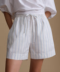 Woman wearing a white blouse with white and blue stripe shorts against a beige background.