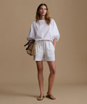 Woman wearing a white blouse with white and blue stripe shorts against a beige background.