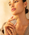 A woman with glowing skin and natural makeup holds a Violet Santal Eau De Parfum bottle with a wooden cap near her face. She wears gold hoop earrings, rings, and a beige sweater while gazing thoughtfully upwards.