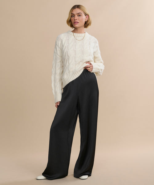 A woman with short blonde hair and a neutral expression stands against a beige background, wearing the Demi Pant, a cream cable-knit sweater, white shoes, and a gold necklace.