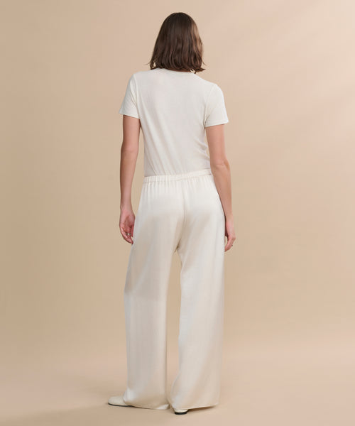 A person with shoulder-length brown hair faces away, wearing a short-sleeve white t-shirt and the Demi Pant against a plain beige background.