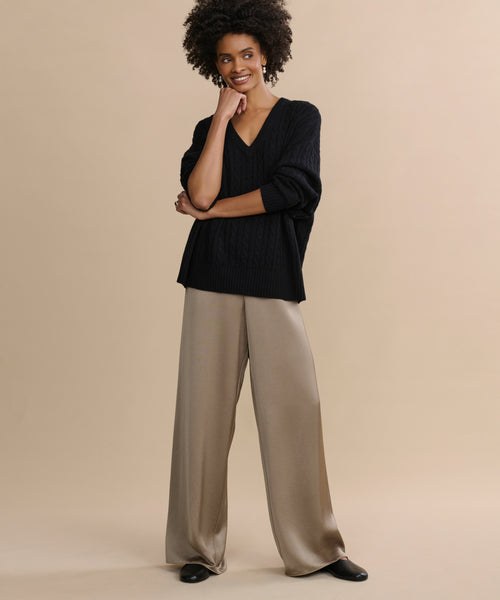 A woman stands against a beige background, smiling as she touches her face while wearing wide-legged taupe satin Demi Pants and a loose black V-neck sweater for an effortlessly flowy look.