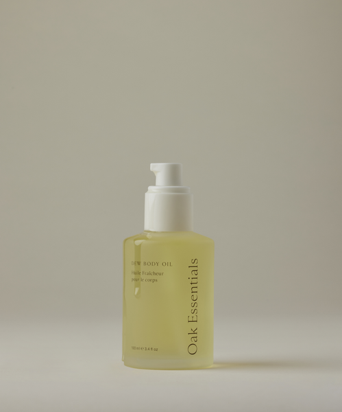 A clear pump bottle of Dew Body Oil, filled with yellow moisturizing oil, stands upright against a plain, light background.