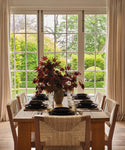 A Dining Table for eight in solid white oak is set with black dishes and a vase of red foliage, positioned before large windows with cream curtains that overlook a garden filled with lush greenery.