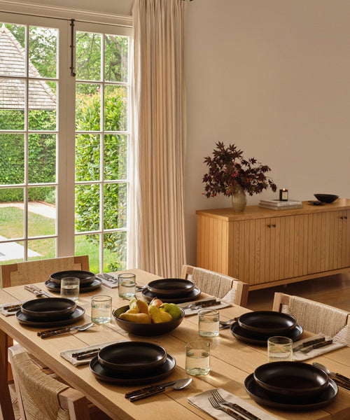 A modern dining room features the Dining Table in solid white oak, set for six with black plates, glasses, and cutlery. A fruit bowl sits at the center; sunlight streams in from large windows and a sideboard with a flower vase is in the background.