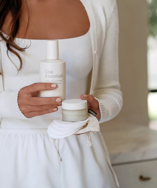 A woman in white holds the Double Cleanse Set—a bottle and small jar—in a white mesh pouch, radiating luxury beauty. The visible labels hint at her secret to a hydrated complexion as she stands indoors.