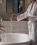 Wearing a white bathrobe, a person sprinkles Dream Bath Soak into a running bathtub in a softly lit bathroom, creating a calming pre-sleep ritual. Only their torso and arm are visible.