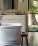 A tranquil bathroom with a white freestanding tub for your Dream Bath Soak, a wooden stool with a candle, a white vanity and mirror, and a large window revealing greenery—an ideal space for relaxing before sleep.
