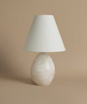 An alabaster base, linen shade table lamp against a beige background.
