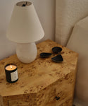 A Droplet Alabaster Table Lamp with a linen shade, a lit candle, and three dark metal accents sit on a wooden nightstand beside a bed with a textured cream headboard.