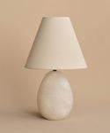 The Droplet Alabaster Table Lamp features an oval stone-like base and a wide linen shade, making it a stylish choice for bedside use against a plain beige background.
