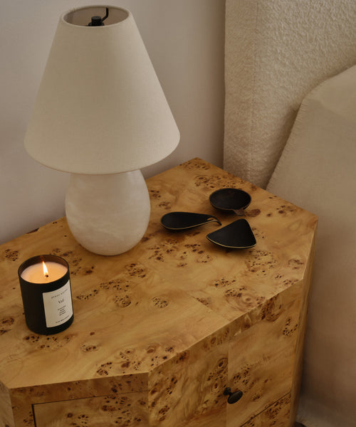 A Droplet Alabaster Table Lamp with a linen shade, a lit candle, and three dark metal accents sit on a wooden nightstand beside a bed with a textured cream headboard.