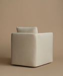 A minimalist cube-shaped upholstered armchair in a warm off-white fabric, set against a solid beige studio background.